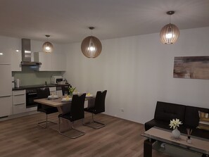 A modern kitchen with white cabinets, a microwave, and a stove. There is a dining area with a table set for two, black chairs, and a vase with flowers. A black sofa and a glass coffee table are in the living area.