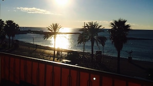 Un balcon offrant une vue sur le coucher de soleil sur la mer, avec des palmiers et une balustrade rouge.