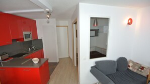 A compact kitchen with red cabinets and a grey sofa in a small living area. - Calysto, 3 *, Heart of Belle-Plagne, on the slopes (La Plagne Tarentaise)