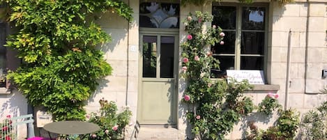 A building with a green door, a window with a sign, a small table with chairs, and a garden with flowers.