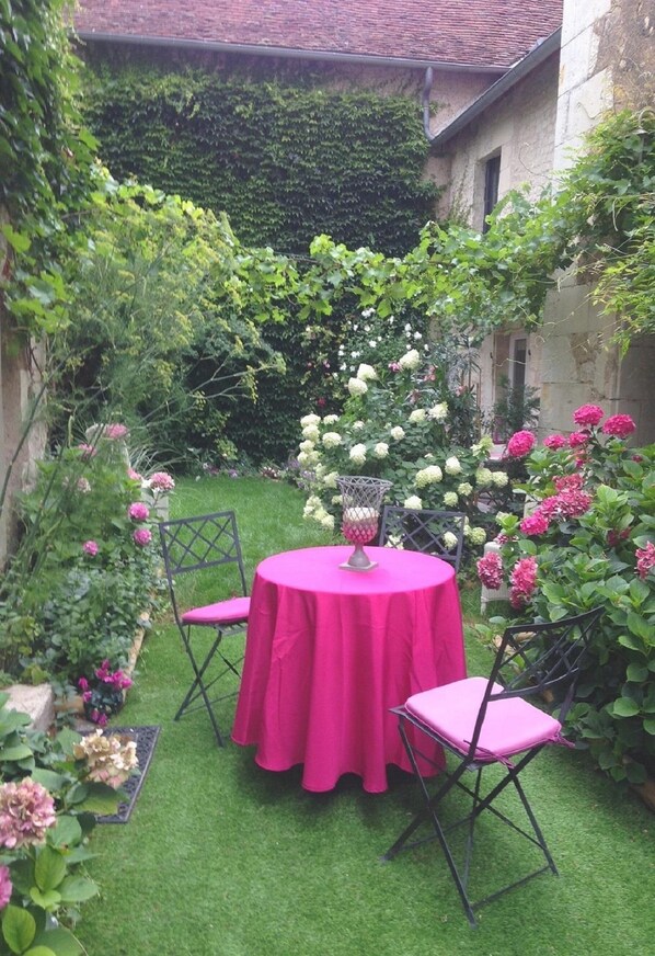A small, enclosed garden with a round table and two chairs, surrounded by lush greenery and flowering plants.