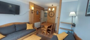 Een knus vertrek met een houten wand, een flatscreen televisie boven een bank, een houten eettafel met stoelen, en een wandlamp.