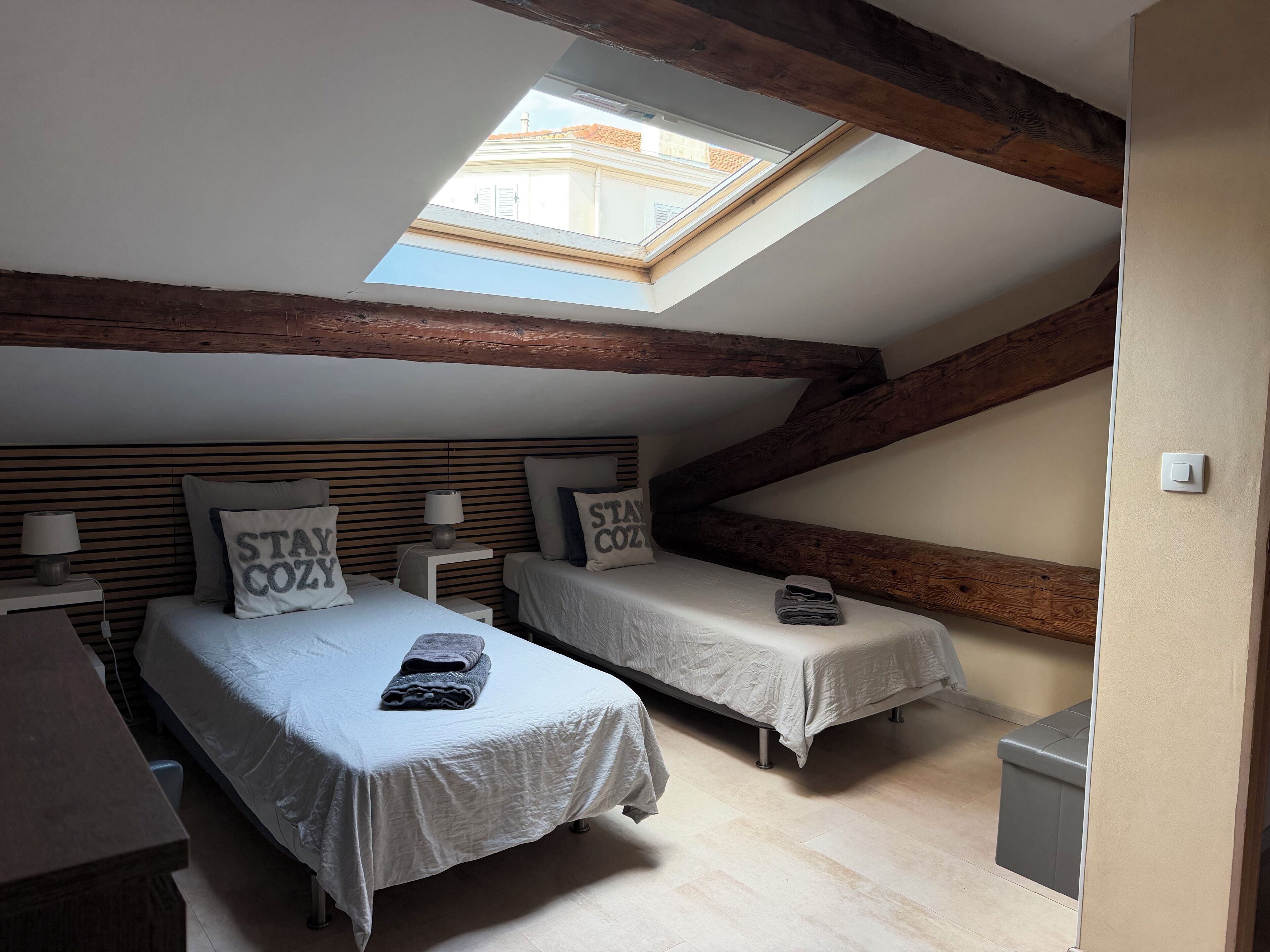 A bedroom with two beds, a skylight, wooden beams, and a small bedside table with a lamp.