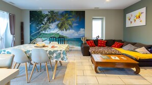 A living room with a beach mural, a dining area with a table and chairs, a sofa with cushions, and a coffee table. - IDEALLY LOCATED GD APARTMENT, PARKING & PRIVATE TERRACE, JACCUZI, QUIET, DOMLOC (Carcassonne)