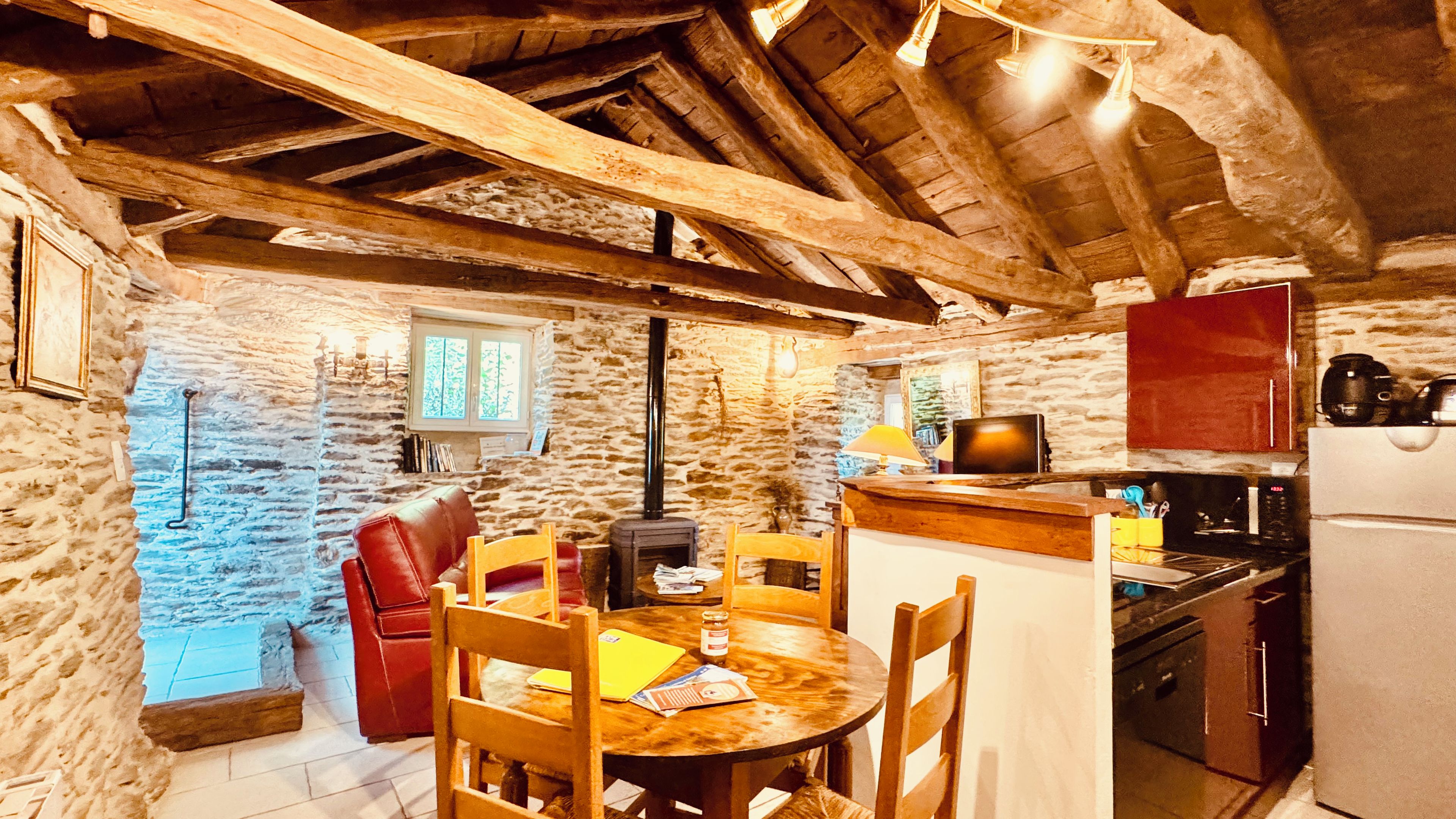 A rustic kitchen with stone walls, wooden beams, a dining table with chairs, a red leather sofa, and a small window.
