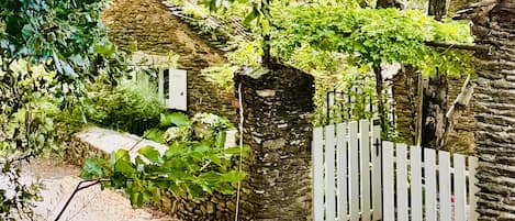 Una cabaña de piedra con valla de piquete blanca, rodeada de exuberante vegetación y un muro de piedra.