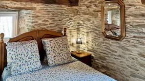 A bedroom with a stone wall, a wooden bed frame, patterned bedding, a bedside lamp, and a mirror. - CASTAGNERE 4* IN THE CÉVENNES: Vacation Rental in Aujac in the Cévennes National Park (Aujac)