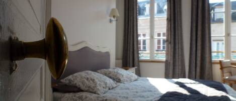 Une chambre à coucher avec un lit, deux lampes de chevet, une fenêtre avec des rideaux et une vue sur les bâtiments à l’extérieur.