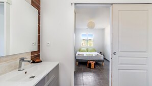 Un bagno moderno con lavandino bianco, specchio e schienale piastrellato. Un corridoio conduce a una camera da letto con letto, finestra e lampada a sospensione.