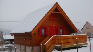 Una baita di legno con tetto imbiancato dalla neve e persiane rosse, con veranda in legno e una scala che conduce all'ingresso.