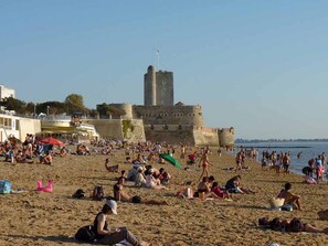 Une plage bondée où les gens se prélassent au soleil et se baignent, une grande tour de pierre en arrière-plan et un ciel dégagé.