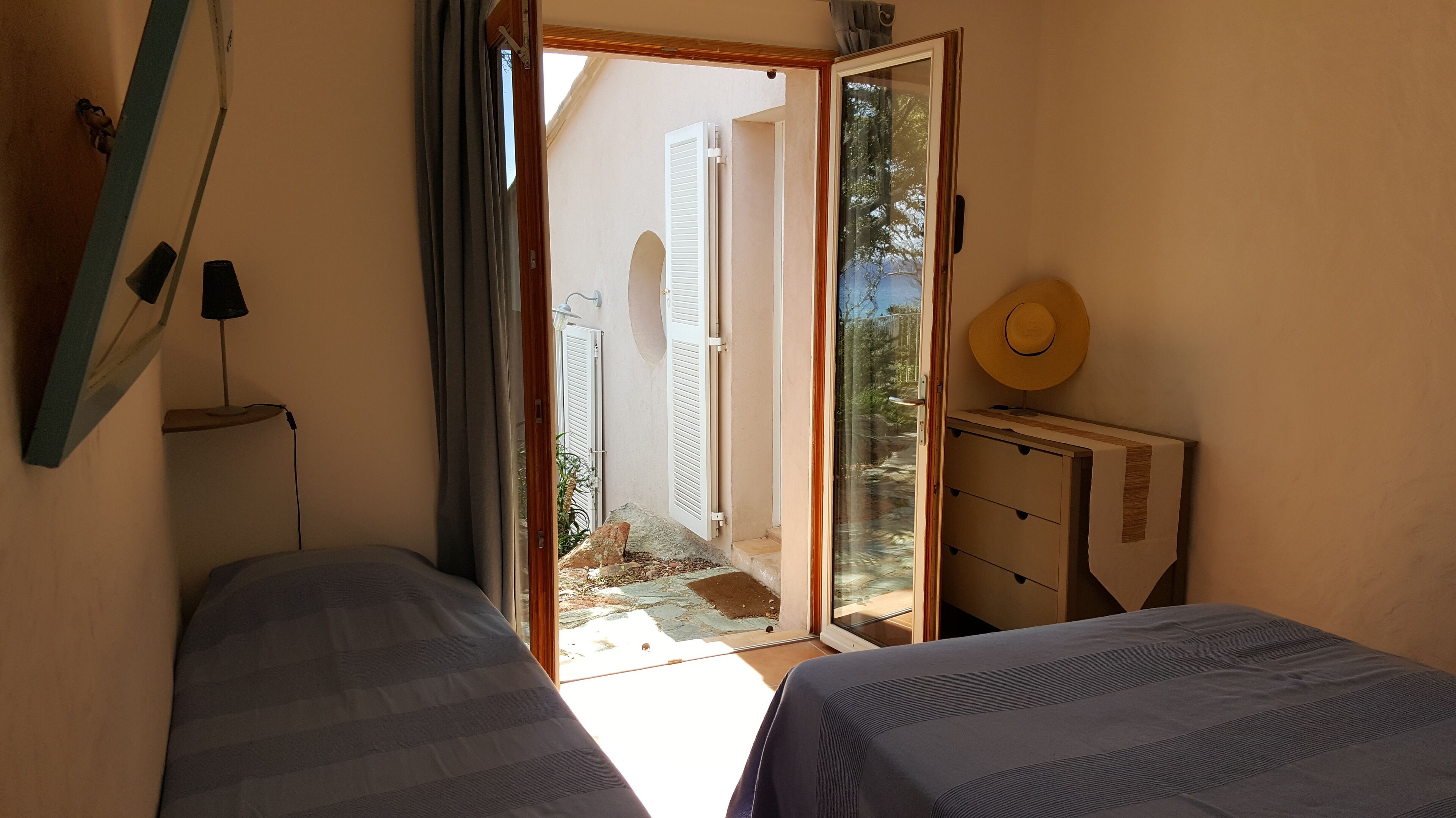 A bedroom with two beds, a desk, a chair, and a wardrobe. There is a window with curtains and a view of the outside.