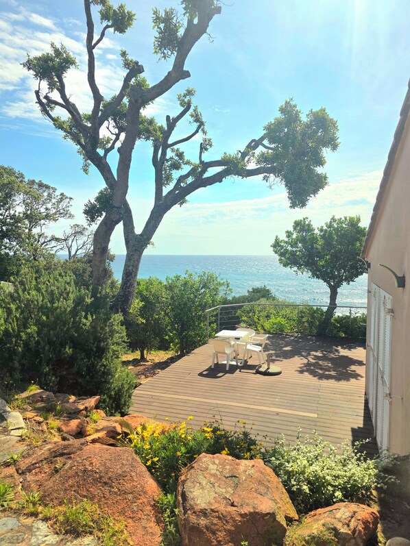 Une terrasse en bois avec des meubles d’extérieur, un grand arbre et une vue sur la mer.