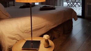 Une chambre d’hôtel avec un grand lit, une table de chevet avec une lampe, une petite table ornée d’un vase de fleurs et une vue sur un espace extérieur enneigé, visible à travers une grande fenêtre.