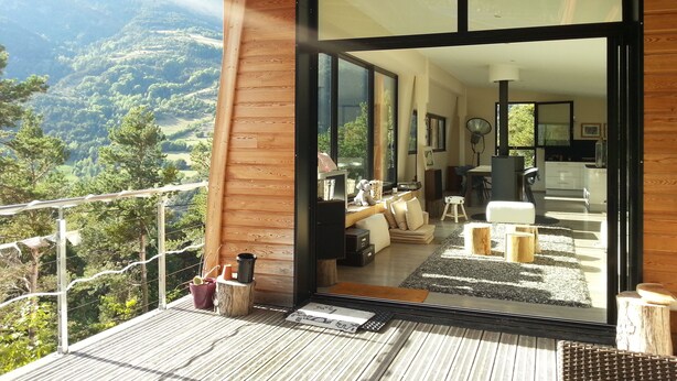 Un balcon en bois moderne offrant une vue sur un paysage montagneux. À l’intérieur, on trouve un espace de vie avec un canapé, une table basse et une table à manger.