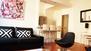 A modern living room with a black sofa, a glass coffee table, and a black chair. There is a white coffee table, a glass of coffee, and a painting on the wall.