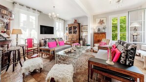 A spacious living room with a large sofa, a glass coffee table, and a chandelier. There are multiple windows providing natural light and a view of greenery outside.