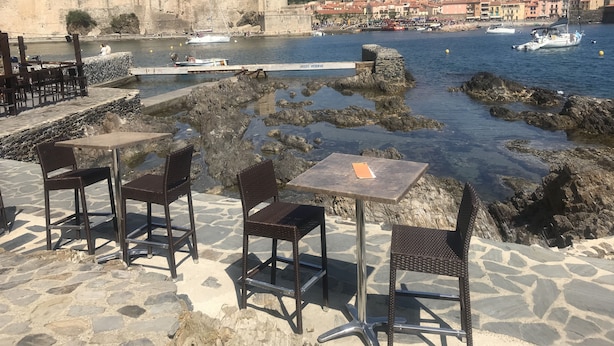 A seaside patio with stone tables and chairs, a view of a historic castle, and boats on the water. - Collioure new air-conditioned apartment with private parking  (Collioure)