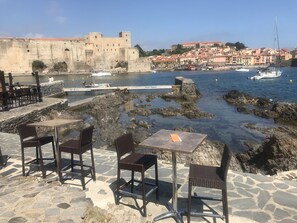 Eine Terrasse am Meer mit Steintischen und -stühlen, Blick auf eine historische Burg und Boote auf dem Wasser.