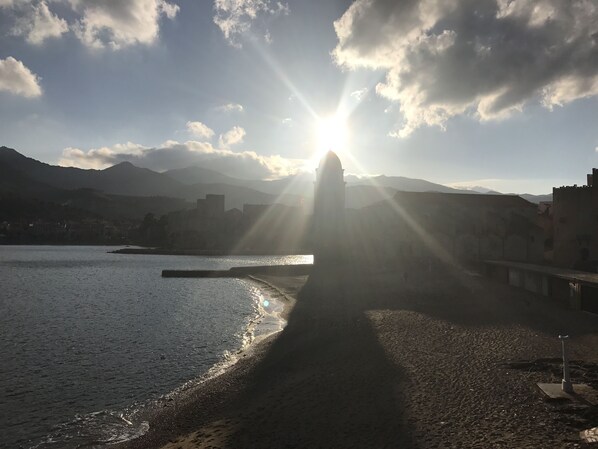 Eine Küstenlandschaft mit einem sonnendurchfluteten Weg, der zu einem Gewässer führt, Bergen im Hintergrund und einem klaren Himmel.