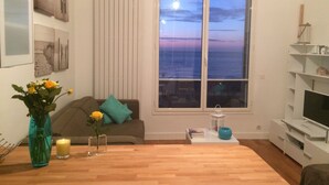 A living room with a wooden floor, a grey sofa with teal cushions, a white radiator, a large window with a view of the sea, and a TV on a white stand. - Trouville-sur-mer feet in the water (Trouville-Sur-Mer)