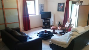Una sala de estar con sofá, sillón y reposapiés, televisión y ventana con cortinas.