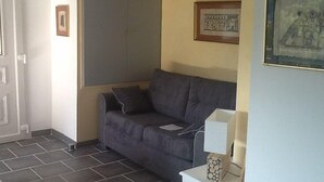 A room with a grey sofa, a white side table with a lamp, a framed picture on the wall, and a door. - Independent apartment in villa, in the pines overlooking the sea. (Ensuès-La-Redonne)