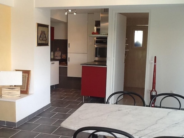 A compact kitchen with white cabinets, a red countertop, and a tiled floor. There is a dining area with a marble table and chairs.