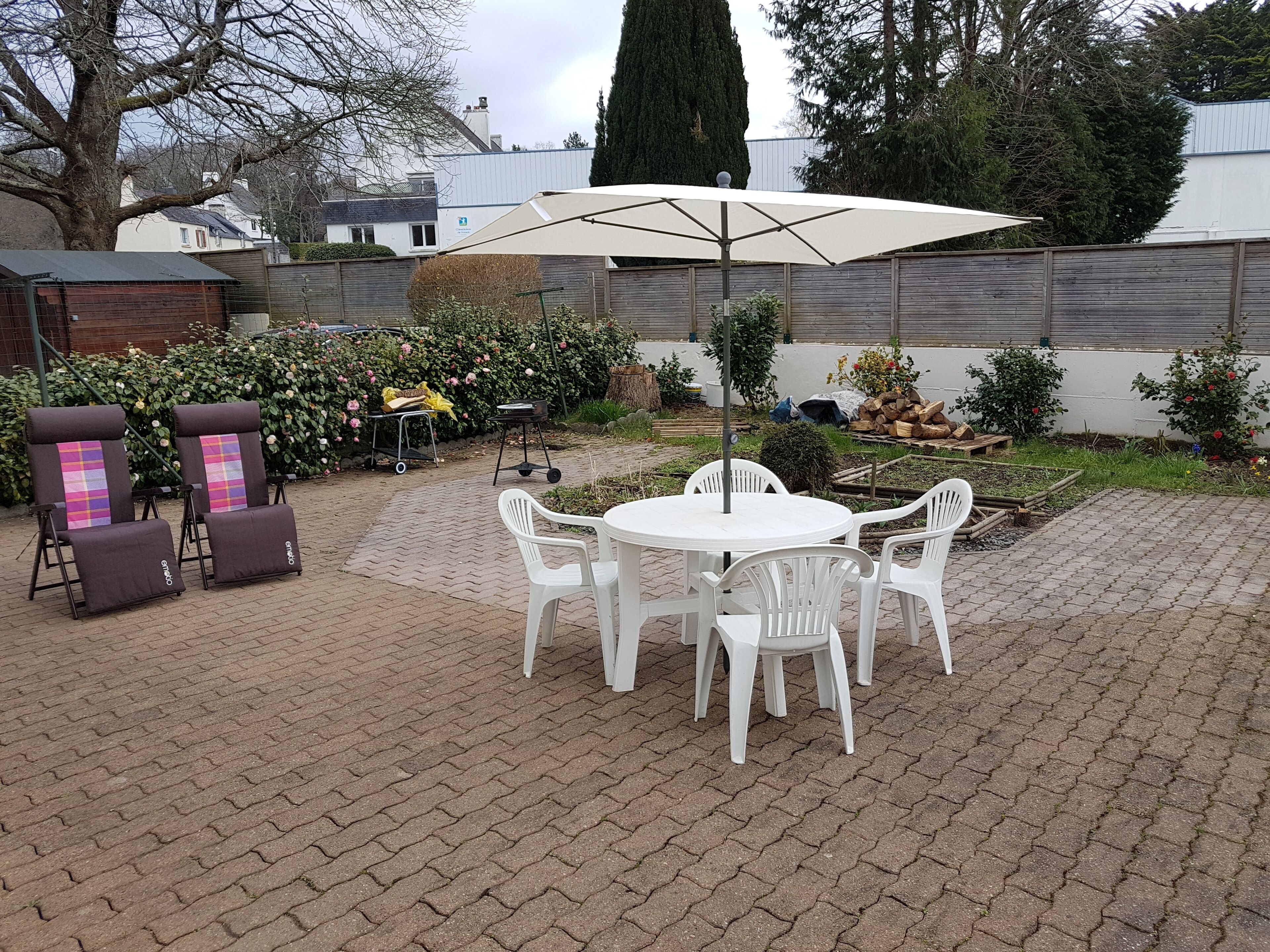 En terrasse med et rundt bord og hvide stole, omgivet af flisegulv, en have med forskellige planter og et træhegn.