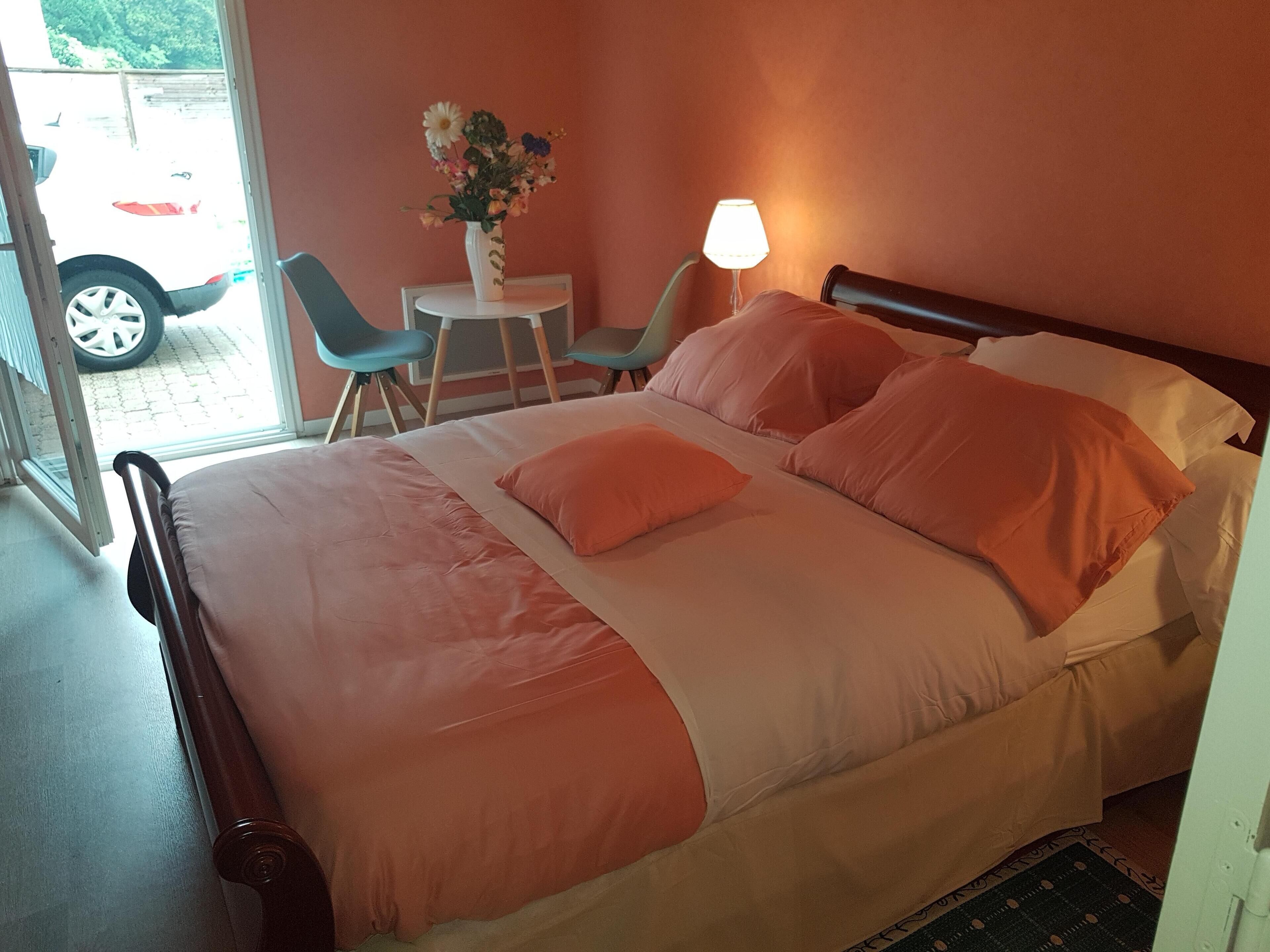 Un lit bien fait, avec une literie orange, une petite table sur laquelle est posé un vase de fleurs et une chaise, dans une pièce d’où l’on peut voir une voiture garée à l’extérieur.