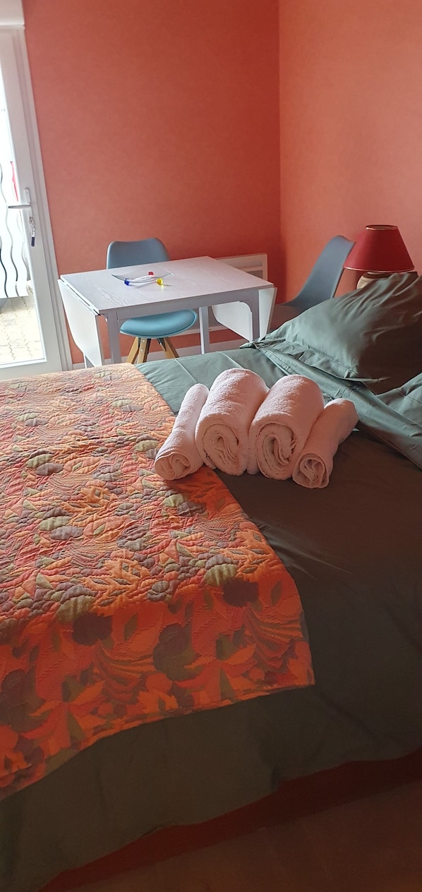 Un lit bien fait, recouvert d’une couverture à motifs floraux et agrémenté de serviettes pliées, une petite table avec une chaise et une lampe rouge.