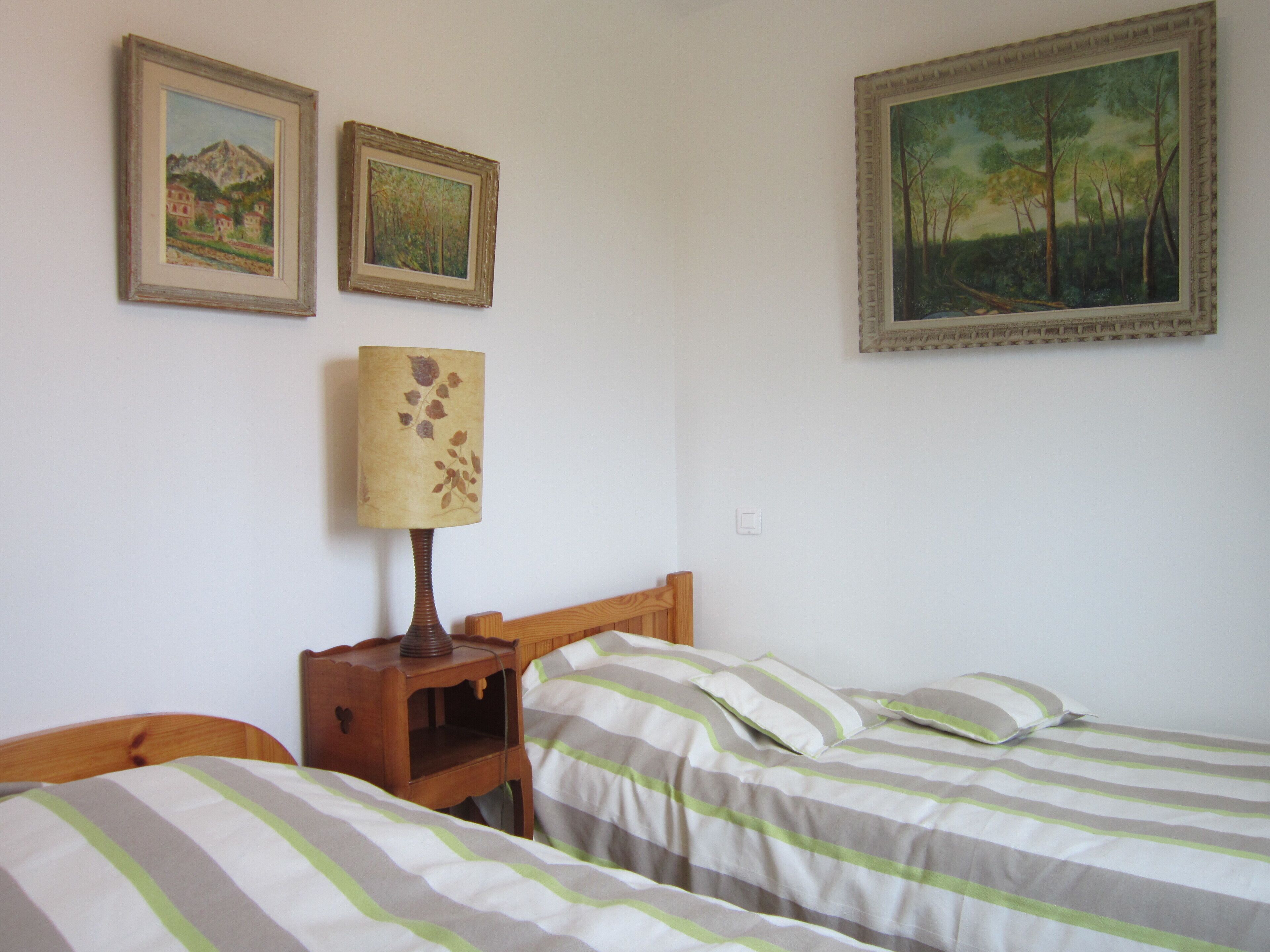 Un dormitorio con dos camas individuales, una mesita de noche con lámpara y dos cuadros en la pared.