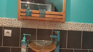 A bathroom with a wooden vanity, a glass bowl sink, a mirror above the sink, and a tiled wall with a pebble-like pattern. - Rental on Amboise (Amboise)