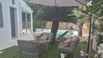 Un jardin avec une pelouse artificielle, deux fauteuils en osier, un grand parasol et une piscine.