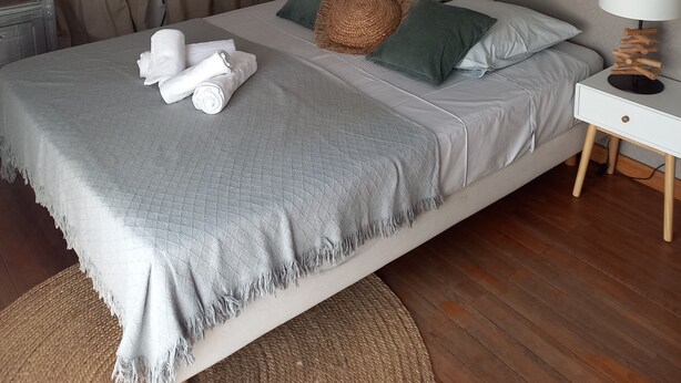 Een net opgemaakt bed met een grijze quilt, twee kussens en een geweven hoofdbord.