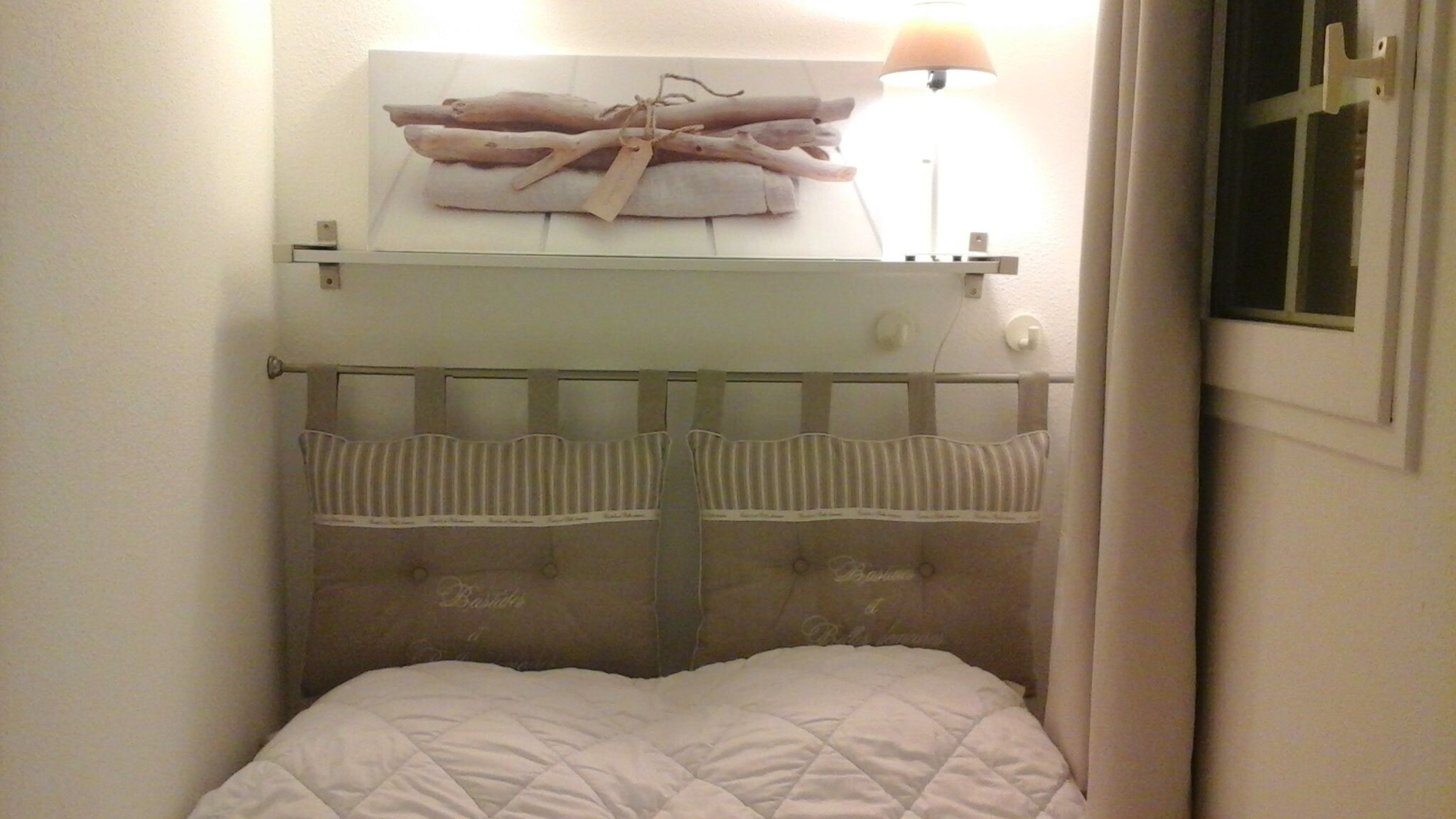 Une petite pièce agréable, dotée d’un lit, d’une table de chevet avec une lampe et d’une étagère murale sur laquelle est posé un objet décoratif.