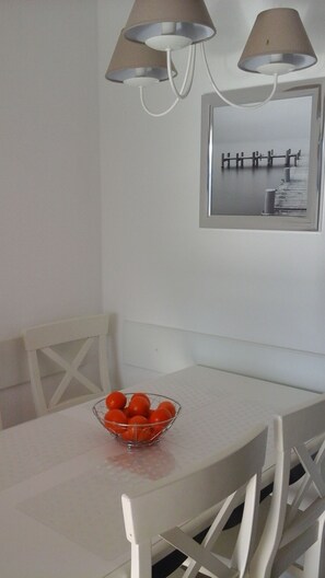 Un coin repas avec une table et des chaises blanches, un saladier rempli d’oranges posé sur la table, et deux lampes fixées au mur.