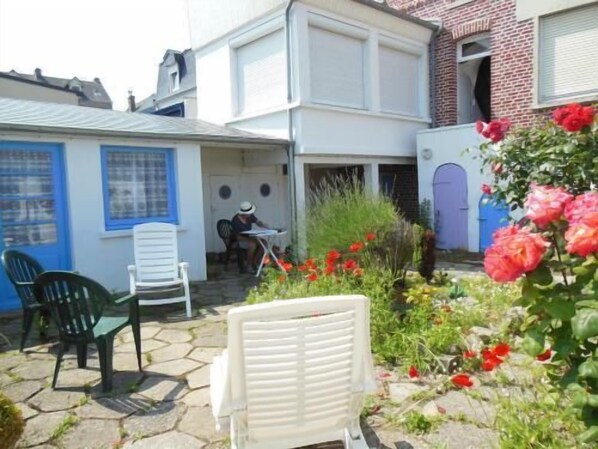 A courtyard with a white chair, a table, and a person sitting. There are red flowers and a green plant in the foreground.
