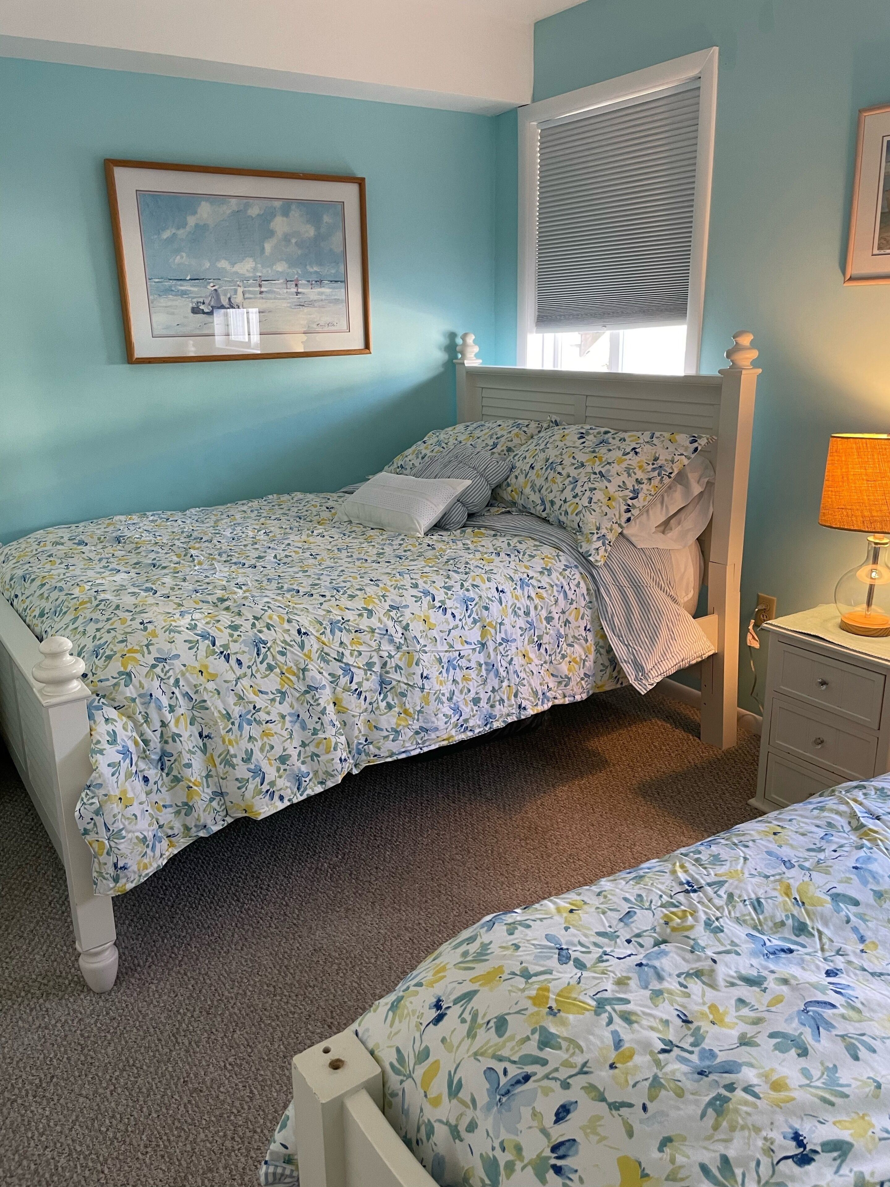 A bedroom with a bed, a nightstand, a lamp, a framed picture on the wall, and a window with blinds.