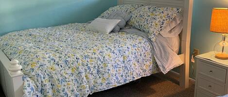 A bedroom with a bed, a nightstand, a lamp, a framed picture on the wall, and a window with blinds.