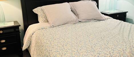 Uma cama com colcha estampada em flor, cabeceira escura e duas mesas de cabeceira com abajures.