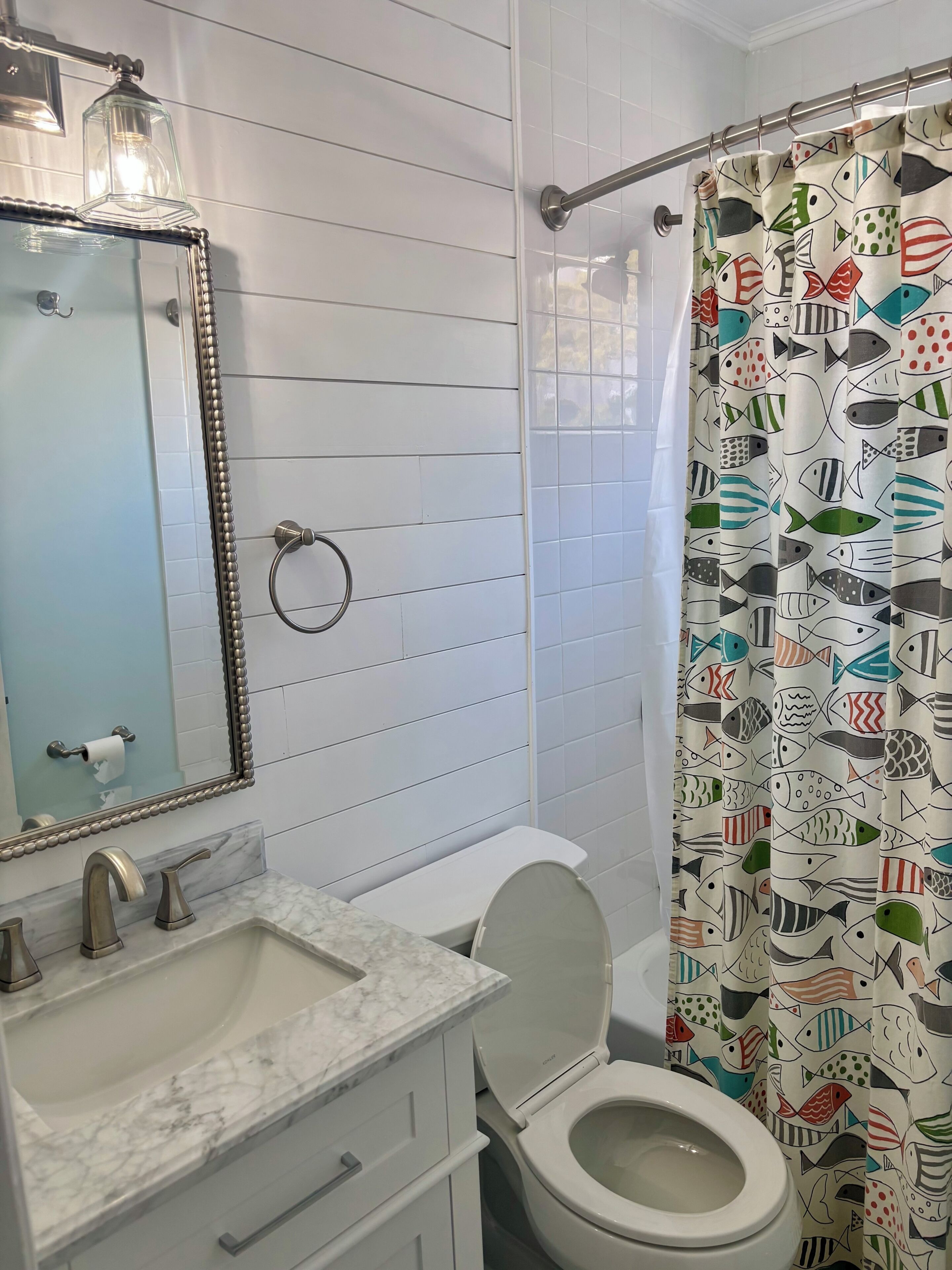 A bathroom with a white sink, mirror, toilet, and shower curtain with fish patterns.