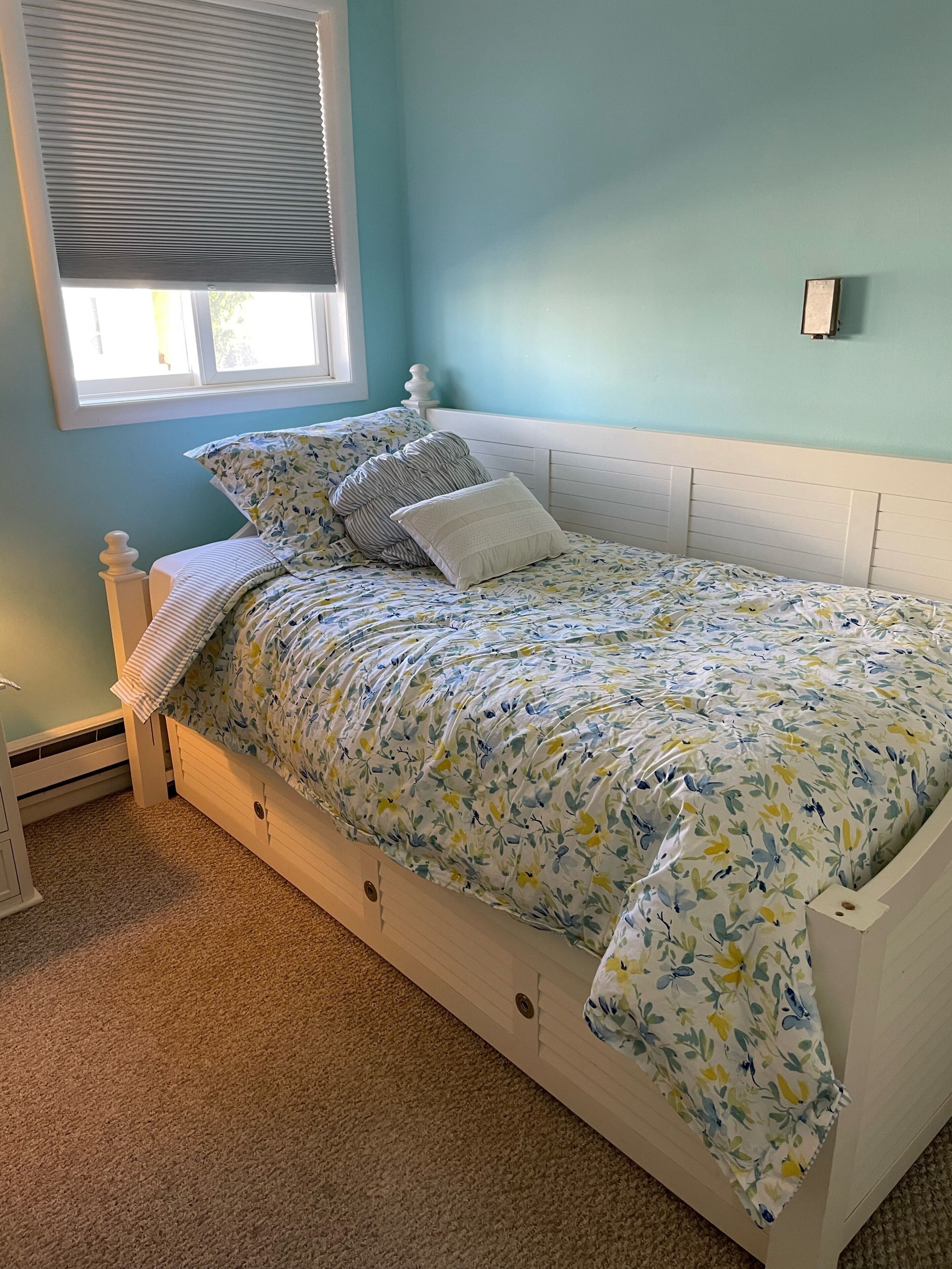 A bedroom with a bed, a window with blinds, a wall clock, and a carpeted floor.
