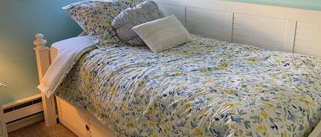 A bedroom with a bed, a window with blinds, a wall clock, and a carpeted floor.