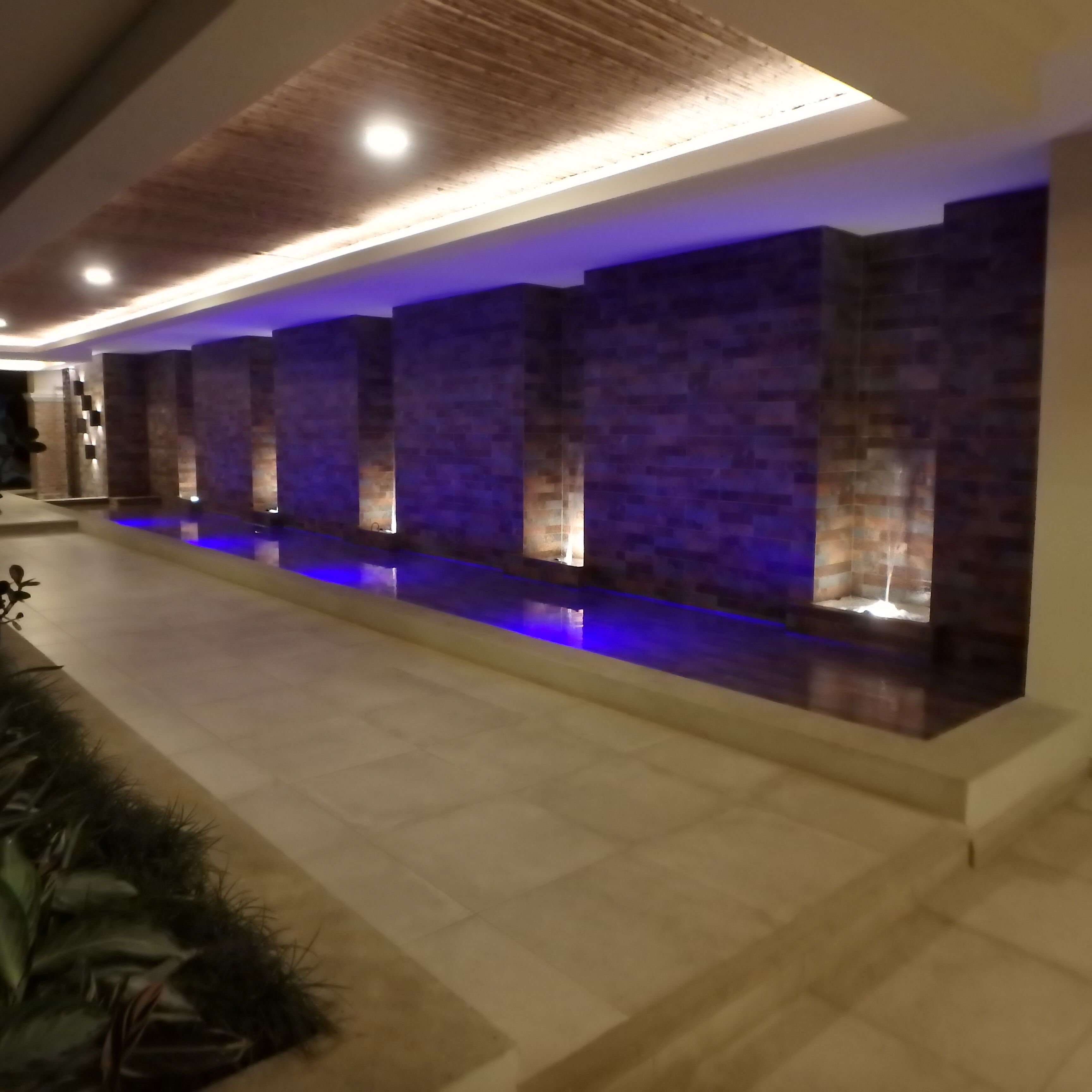 A modern indoor pool area with a long, narrow pool, tiled walls, and recessed lighting.