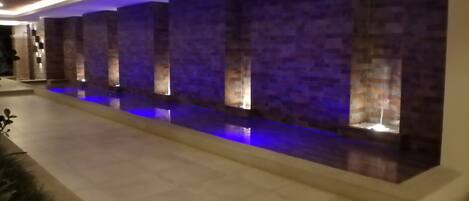 A modern indoor pool area with a long, narrow pool, tiled walls, and recessed lighting.