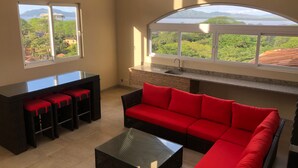 Ein modernes Wohnzimmer mit einer Couch, einem Couchtisch und einem Essbereich mit Tisch und Stühlen. Große Fenster bieten Ausblick auf eine Berglandschaft.