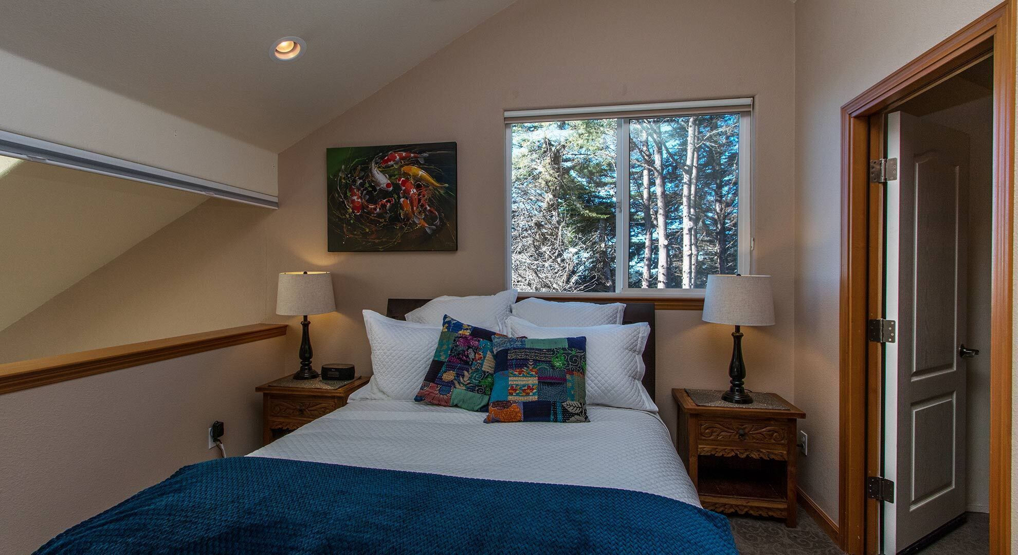 A bedroom with a large bed, two nightstands, a window with a forest view, and a painting on the wall.
