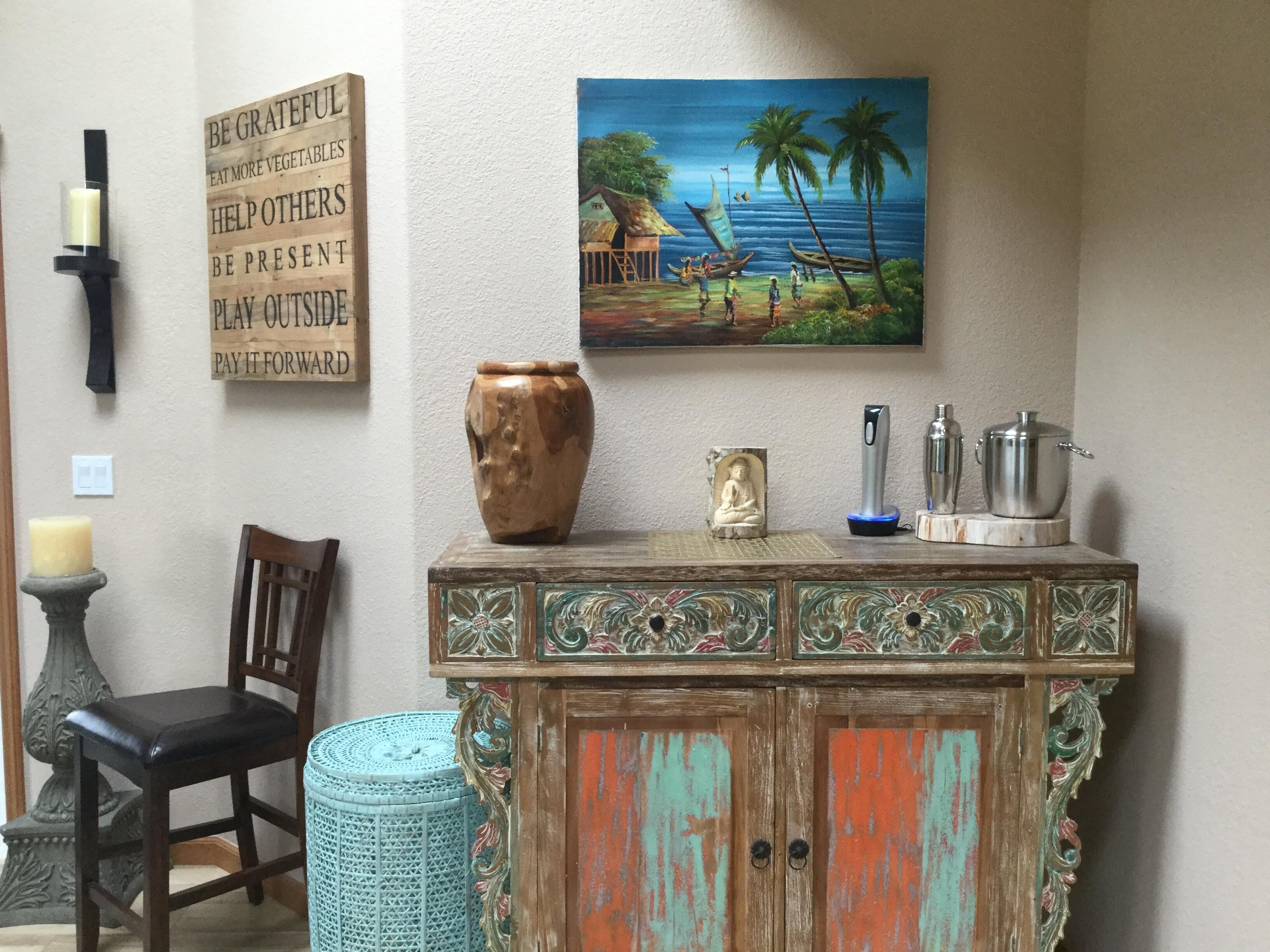 A room with a wooden cabinet, a painting of a tropical scene, a candle, and a chair.