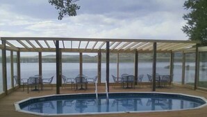 A covered outdoor swimming pool with wooden decking and glass panels, surrounded by clear chairs and tables. - Private Lodge-Mtn Lake, Pool,Hot Tub, Affordable Family Reunion Sleeps 20+ (Westcliffe)
